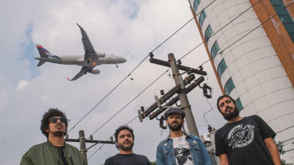 Imagem dos quatro integrantes da banda em frente a um prédio e um avião sobrevoando o céu atrás