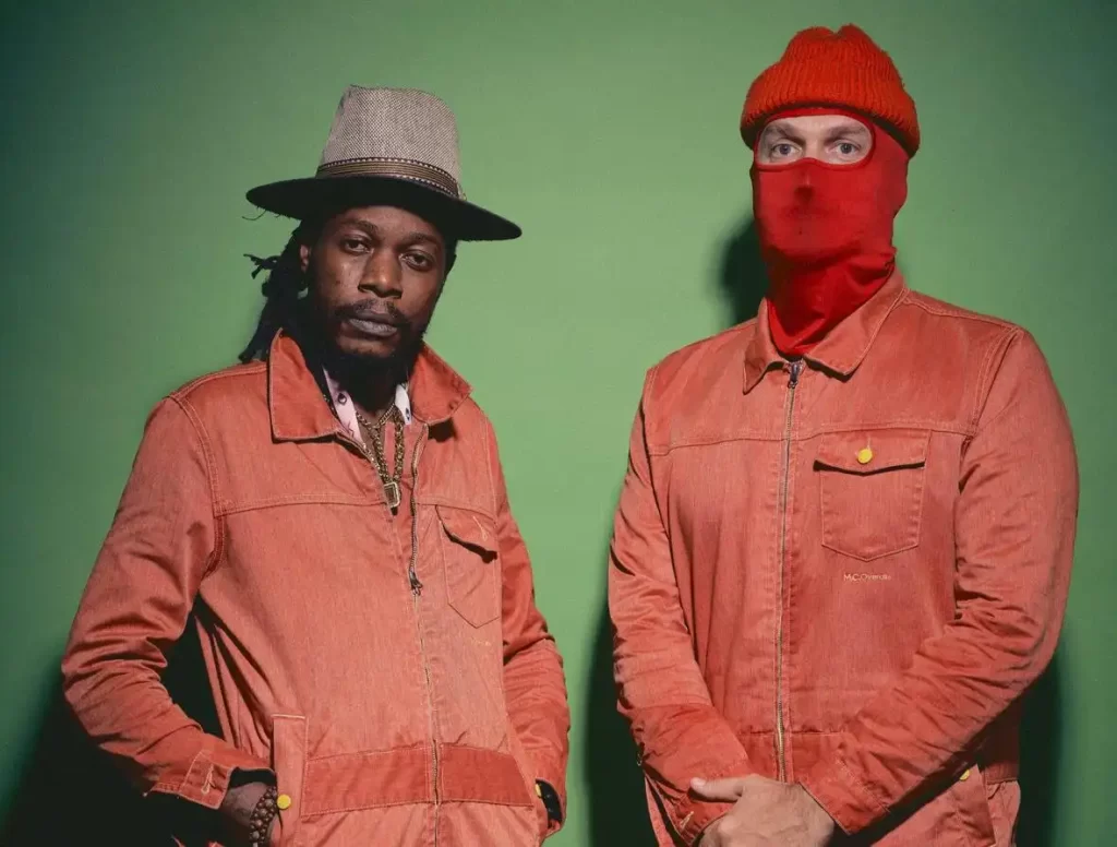 Imagem de dois integrantes da banda Kokoko! vestidos de laranja em um fundo verde